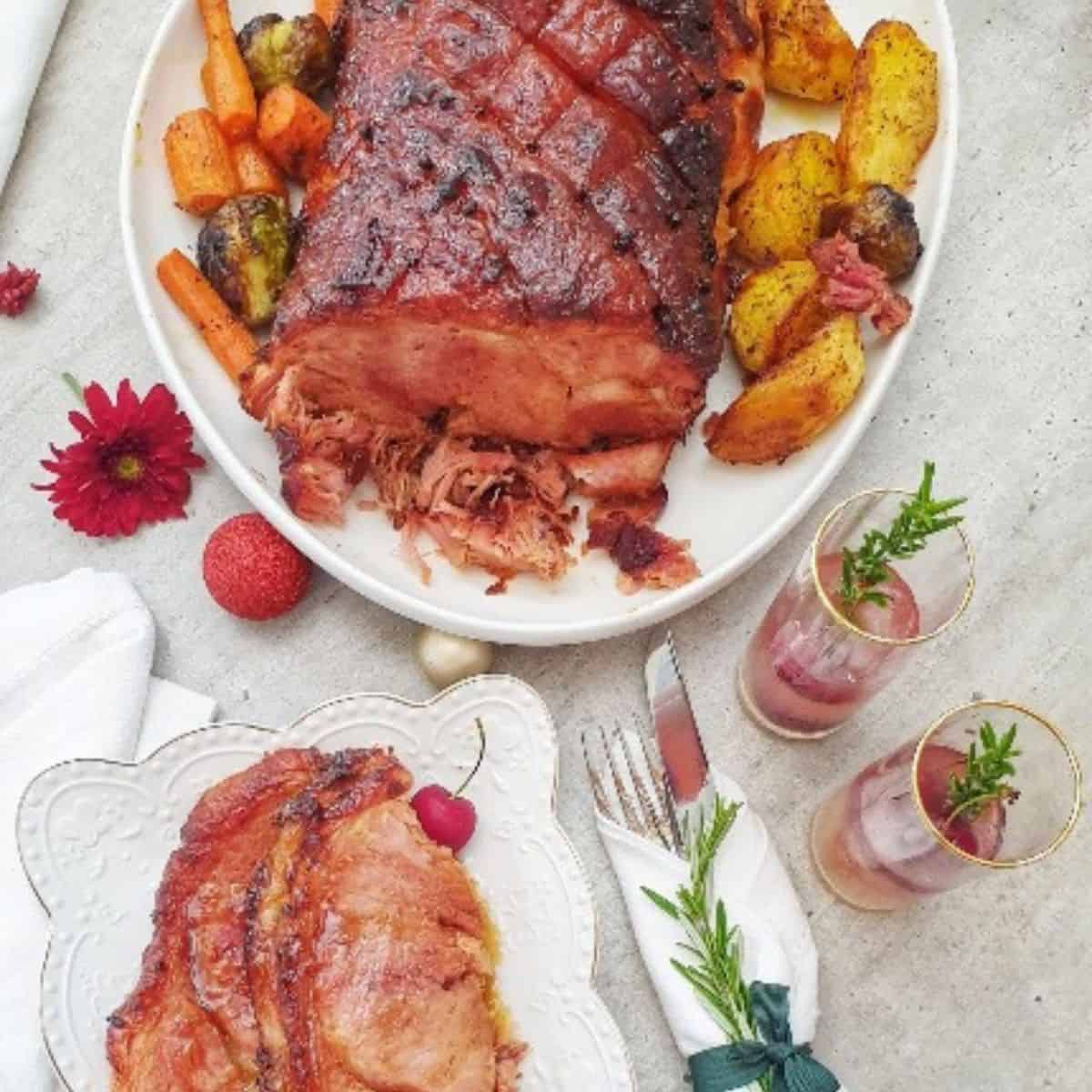 On a white table is cooked and glazed gammon on a white serving platter, with roasted veggies – carrots, brussel sprouts and potatoes. The gammon fat is brown and glazed. The smaller second platter has sliced gammon, veg and cherries around. Cutlery wrapped in a white serviette with fresh rosemary for décor. Tall glassed with red color drinks in and sprigs of fresh rosemary. Xmas décor around the table.