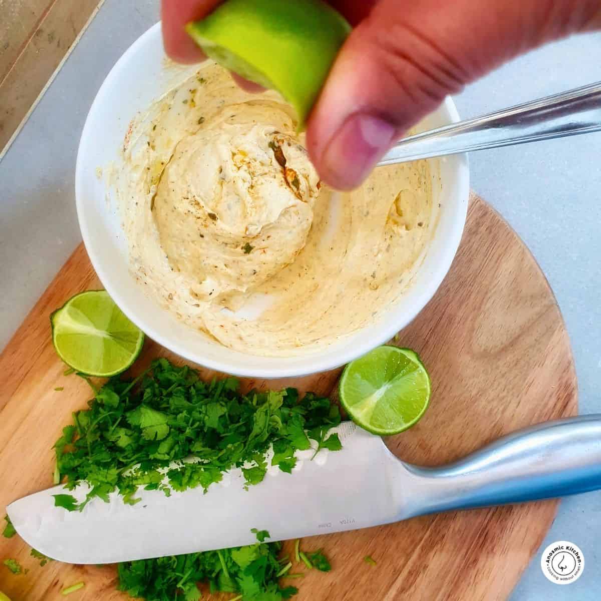 lime being squeezed into a mascarpone cheese dip with chopped fresh herbs a kitchen knife on a board.