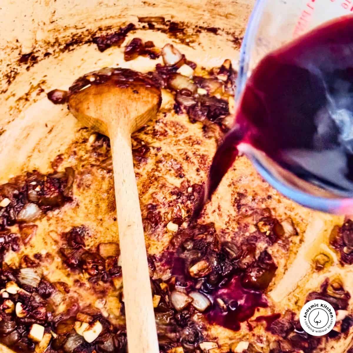 red wine being poured into a pot with fried onions. Wooden spoon in pot