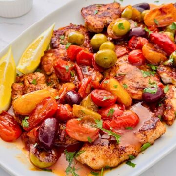 pan-fried yellowtail fish recipe with cherry tomatoes and olives, lemon wedges and.a sauce on a platter, wtih small bowls of freshly chopped herbs around.