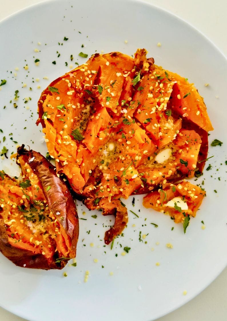cooked air fryer sweet pototo halved on a platee with butter, hemp seeds and parsley for garnish.