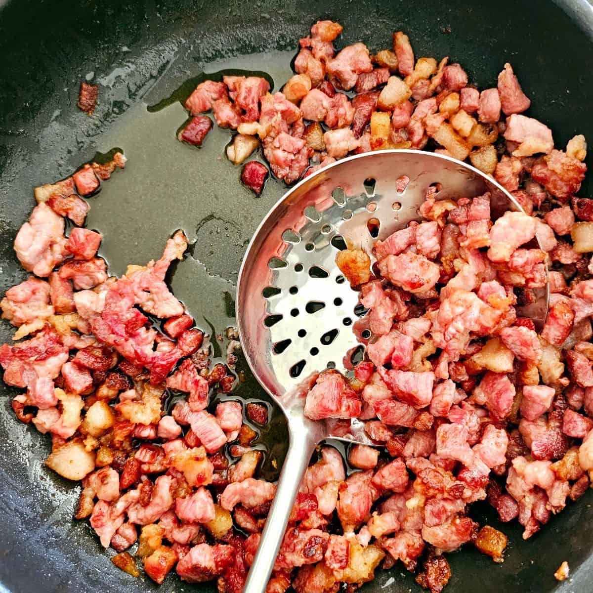 Crispy fried bacon pieces in a skillet with a large spoon to scoop and drain excess oil for creamy bacon pasta.