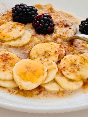 cooked oats with sliced banana, fresh blackberries, cinnamon, honey and a spoon in a white small bowl