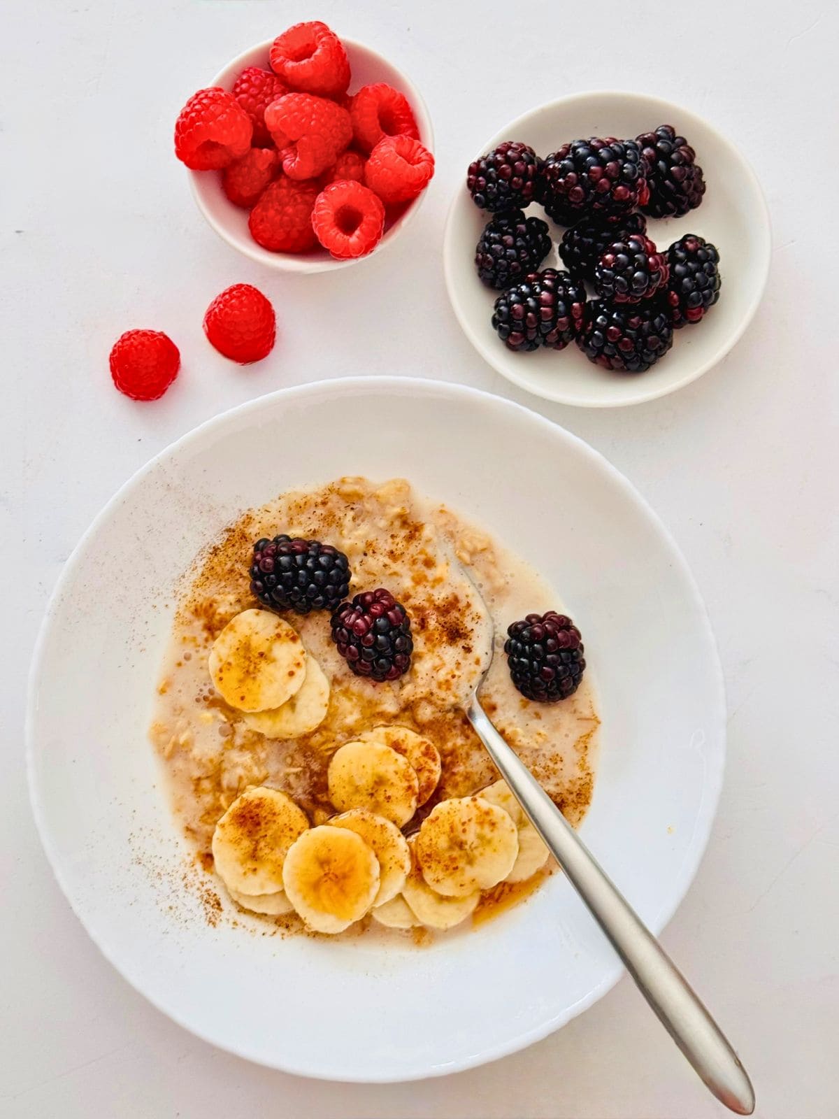 cooked oats with fresh fruit in a small white bowl and fresh berries around it. 