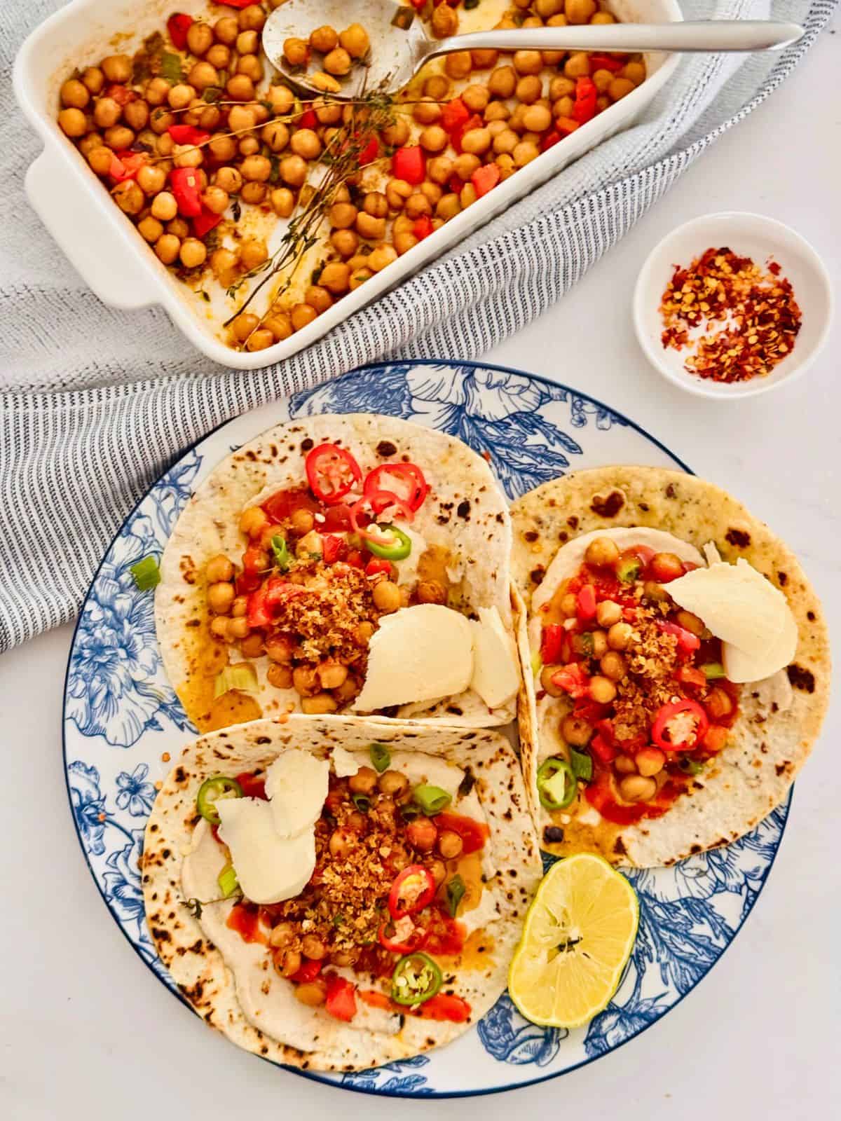 air fryer roasted chickpeas on small wraps on a blue floral plate with fresh lemon. Dried pepper flakes in a small bowl and a roasting dish with chickpeas and peppers  all on a table. 