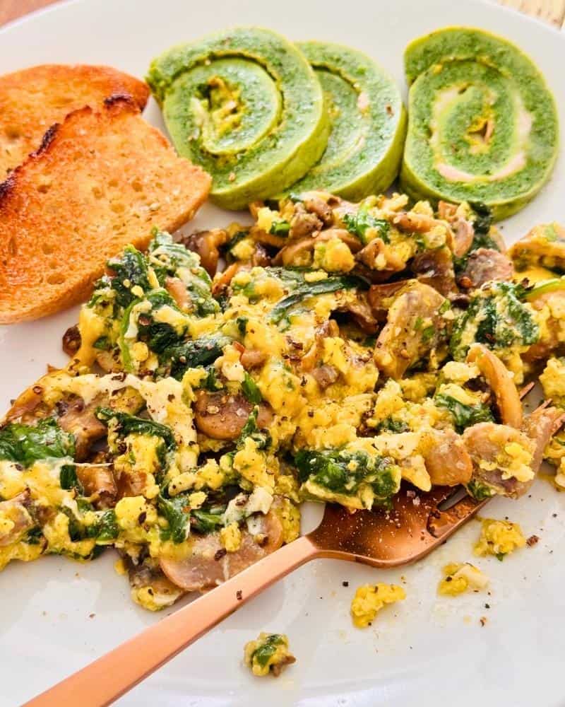 Scrambled eggs with spinach and mushrooms on a white plate, served with toast and slices of rolled spinach omelette.