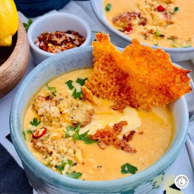 Air fryer butternut squash soup topped with roasted seeds, herbs, and a lacy cheese crisp, with chili flakes and lemon in the background.