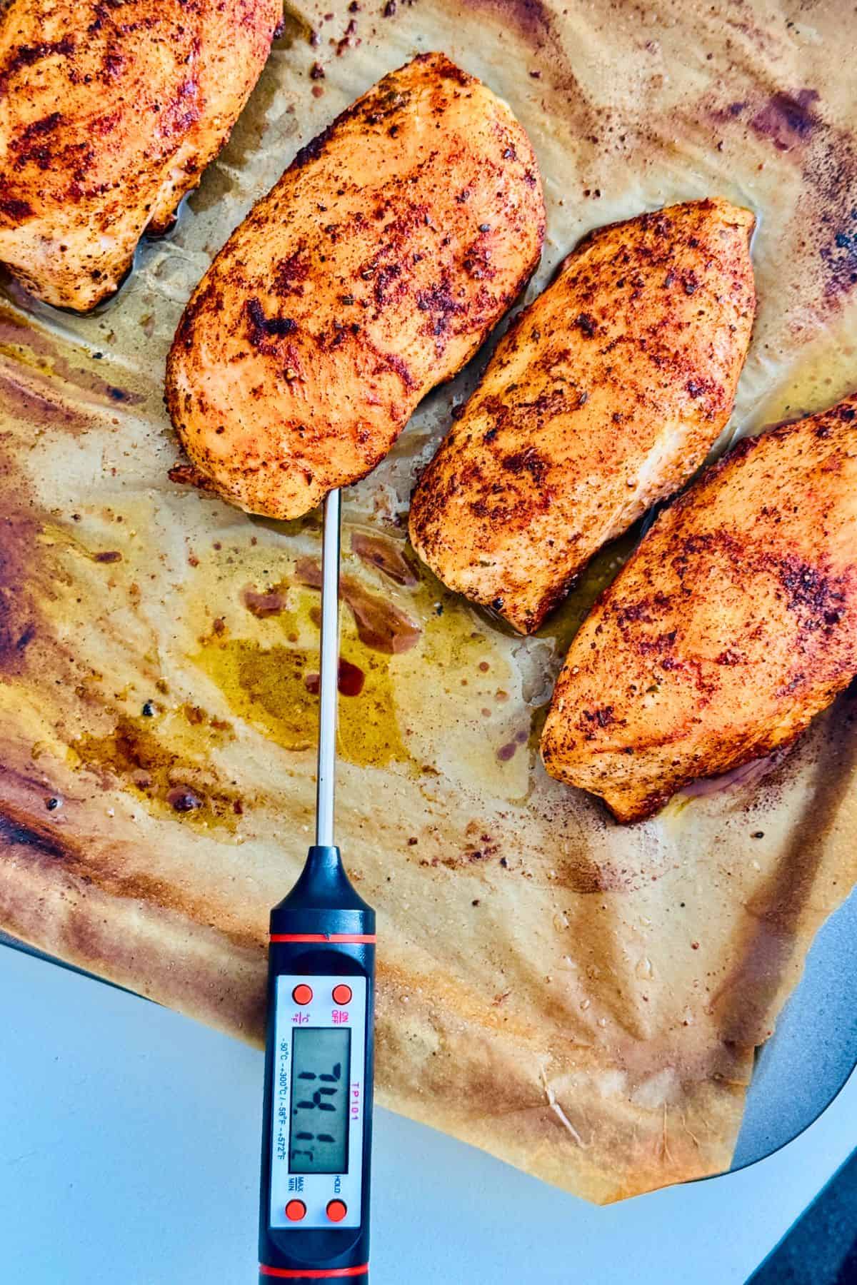 Oven-baked chicken fillets on parchment paper with an instant-read thermometer inserted showing cooked internal temperature