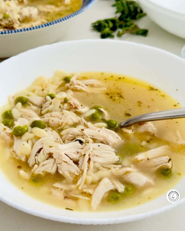 Simple chicken soup in a white bowl, topped with black pepper and olive oil, spoon beside it, fresh oregano nearby.