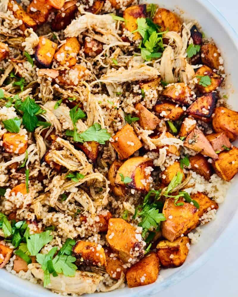 A colorful bowl filled with couscous, roasted sweet potato cubes, and shredded chicken, garnished with herbs and served with a drizzle of sauce.