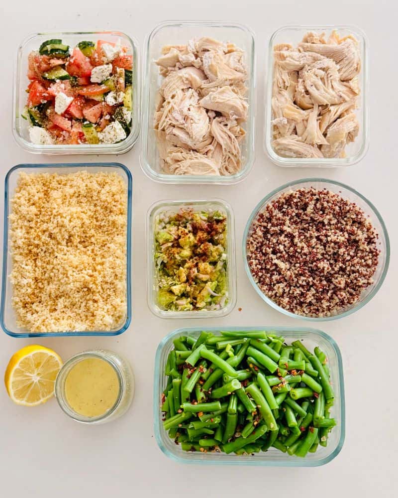 Meal prep containers with cooked chicken, green beans, couscous, quinoa, guacamole, tomato and feta salad, and fresh lemon on a white table.