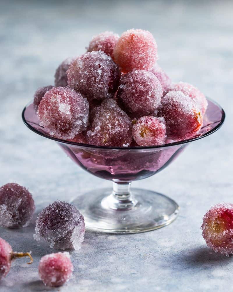 Frozen red grapes covered in frost served in a small glass bowl, with a few loose grapes scattered around.