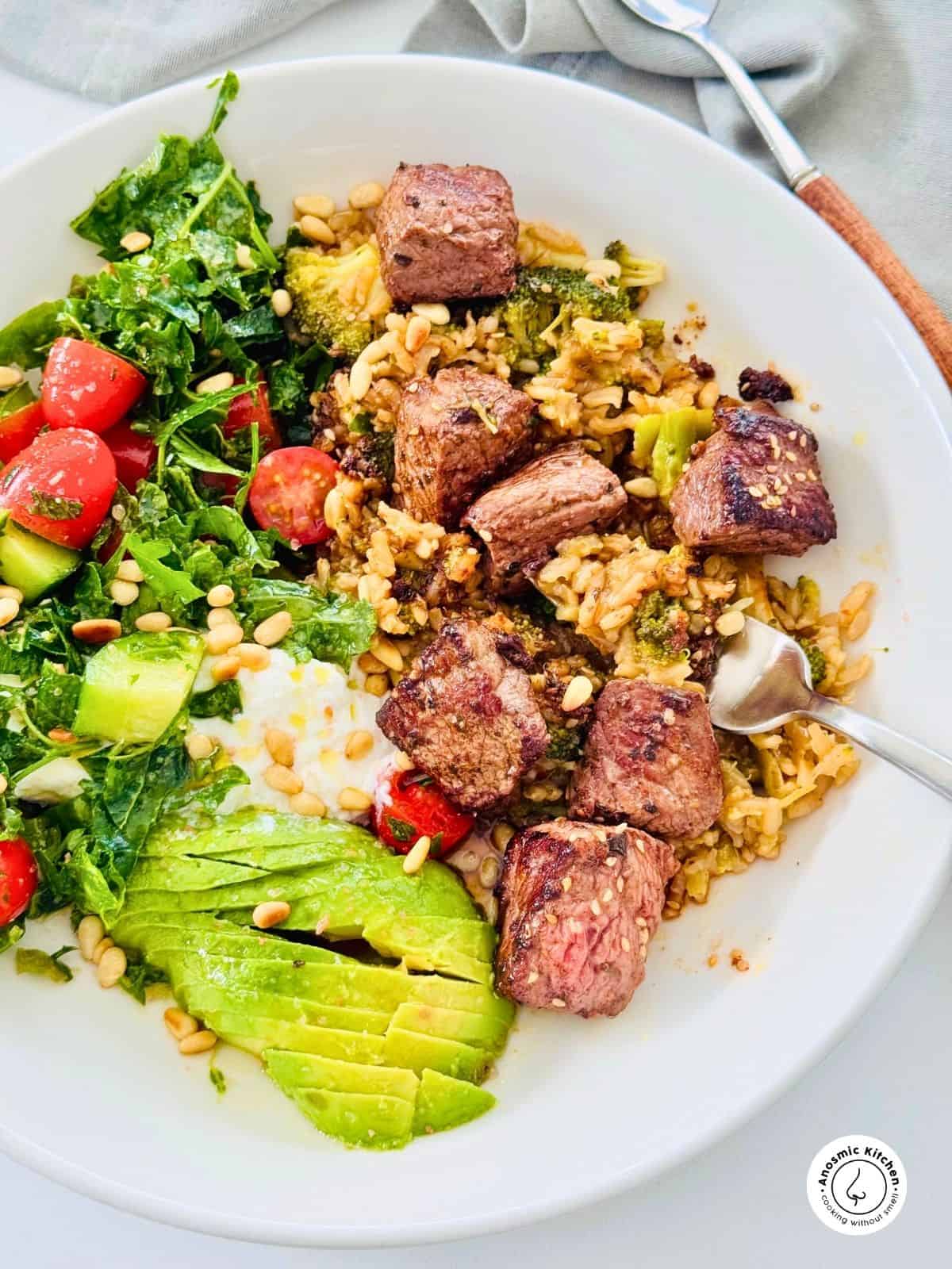 Healthy lunch bowl with brown rice, seared ostrich cubes, broccoli, avocado, kale salad, and yogurt on a white plate.