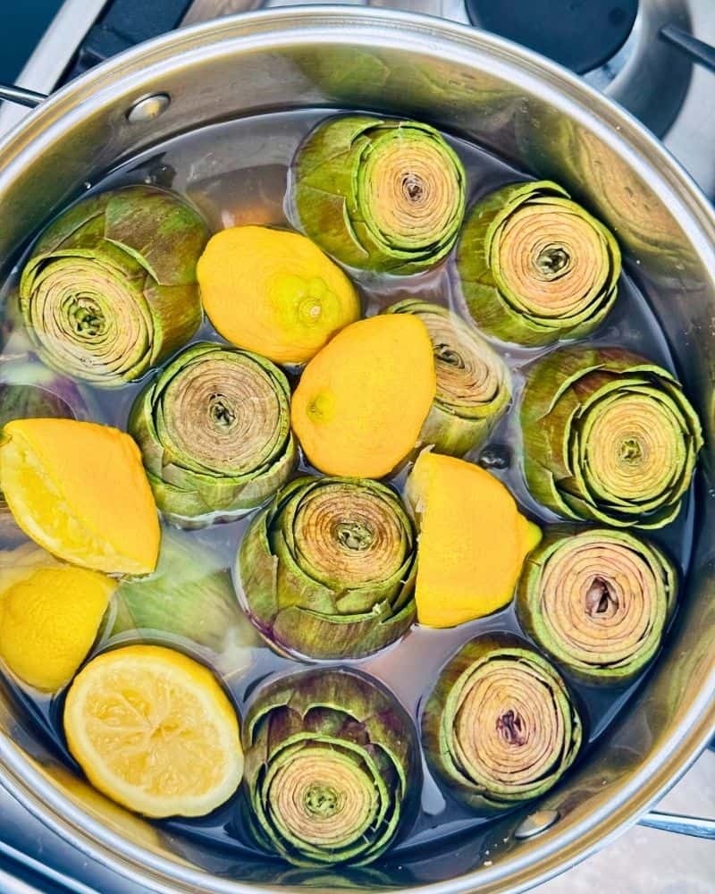 Halved artichokes cooking cut side up in a pot of water with lemon halves
