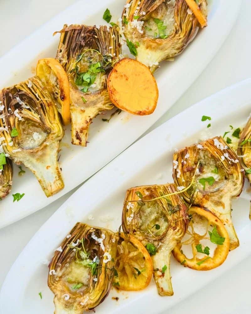 Sautéed artichoke starters on white platters with browned lemon slices, parmesan, sea salt, and fresh thyme.