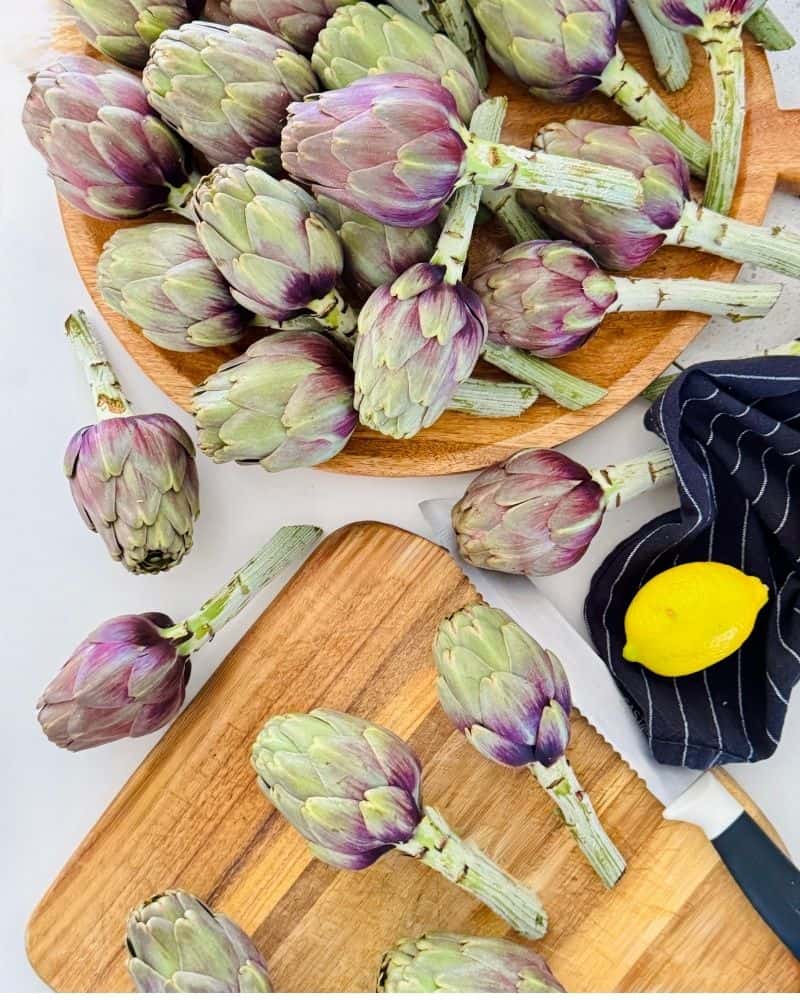 Fresh artichokes on wooden boards with a black and white checked dish towel, serrated knife, and lemon.