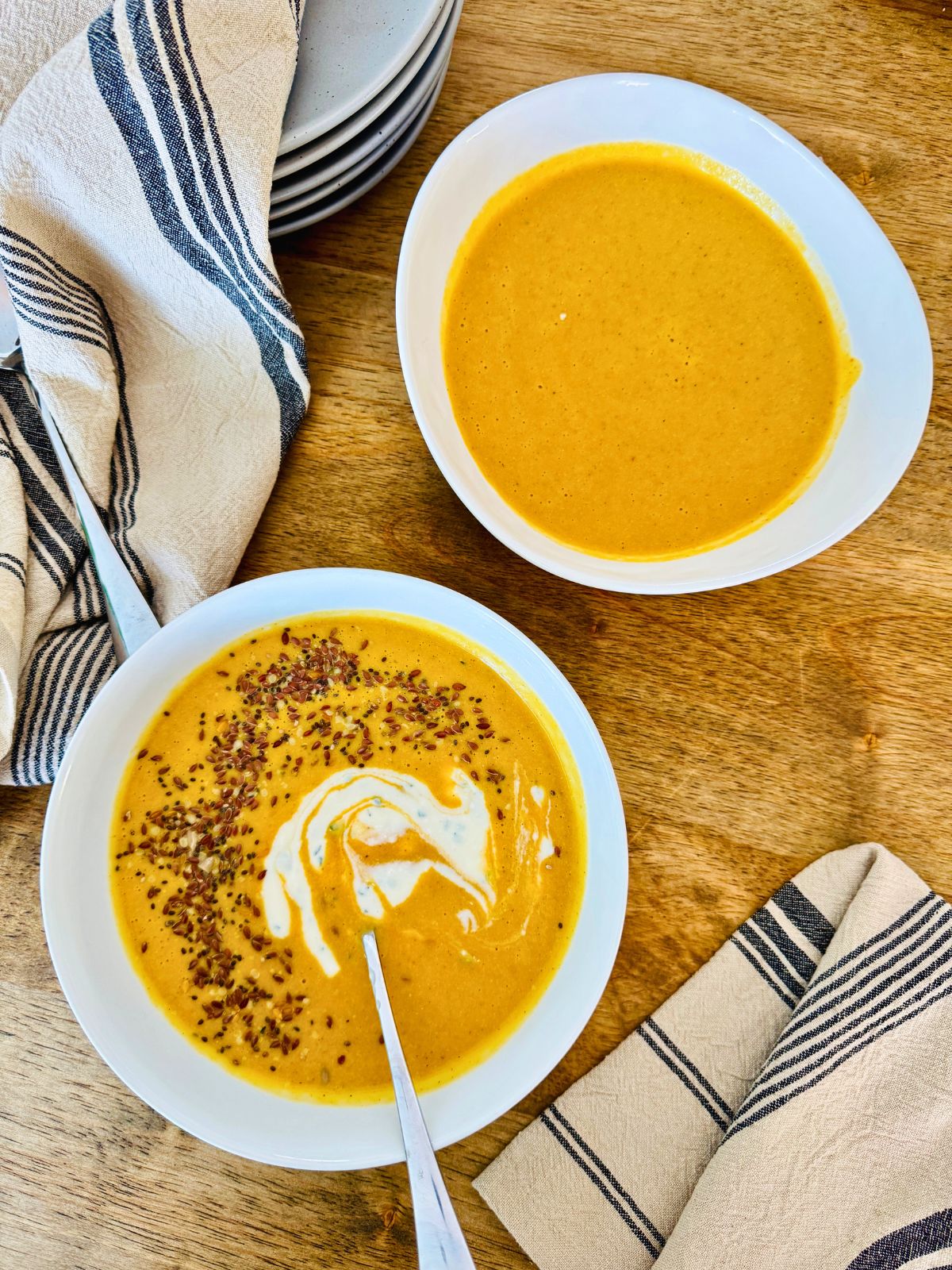 veggie soup in 2 white bowls and spoons and serviettes. One soup bowl has cream and seeds in it.