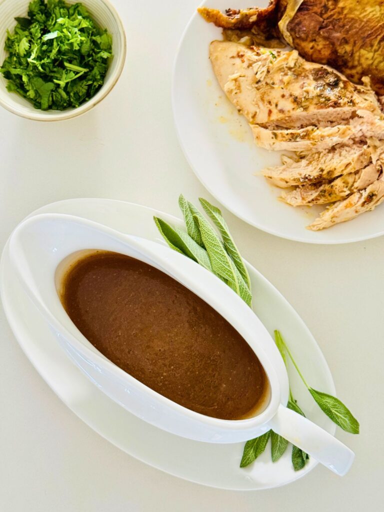 chicken gravy in a white gravy bowl with sage on the side and cooked chicken breast next to it with a small bowl of chopped herbs on the other side.