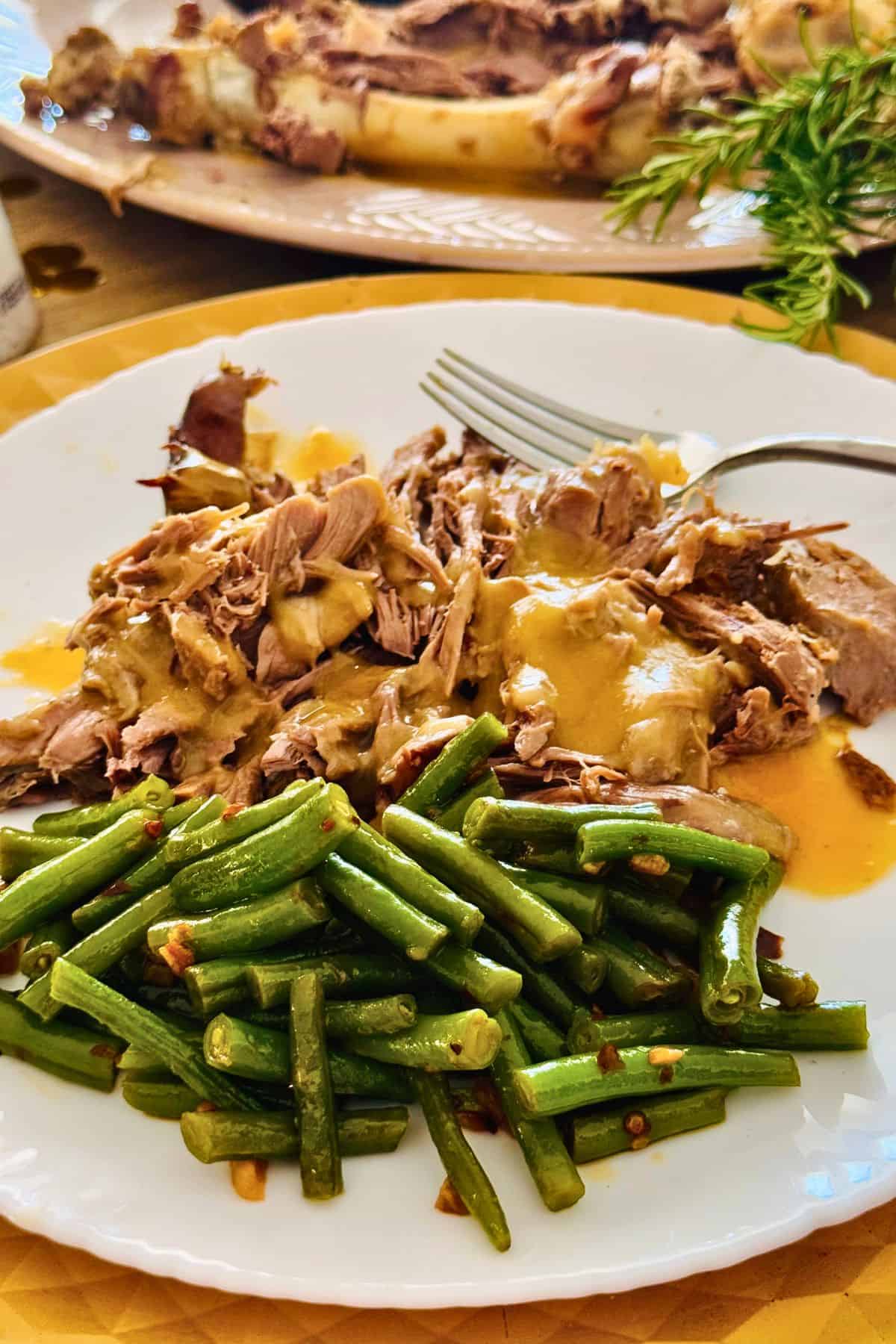 Fall-apart roasted leg of lamb served on a white plate with golden gravy and a side of green beans.