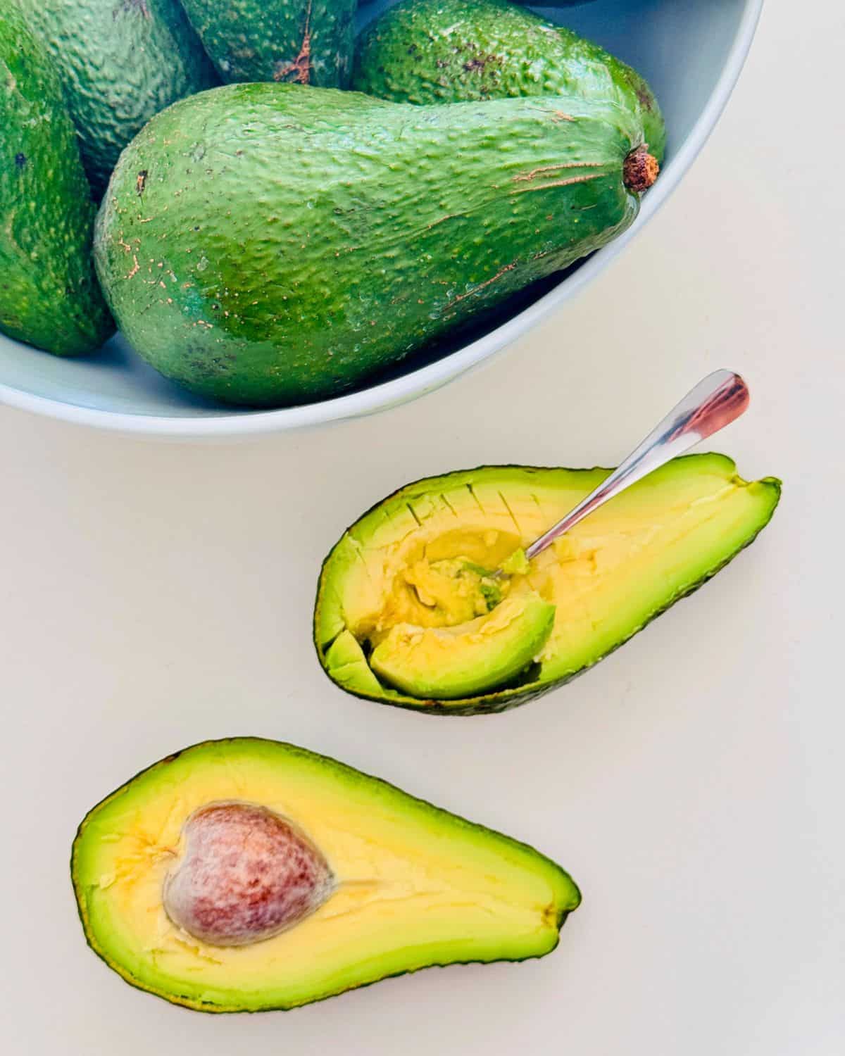large avocados in a white bowl, one sliced in half showing the creamy texture with a spoon in the one cut half.