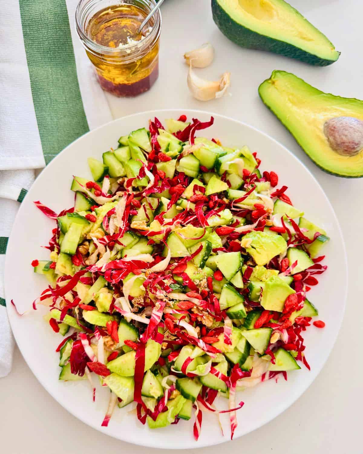 radicchio cucumber and avocado chopped salad on a white plate. Halved avocado, garlic and a mason jar with a vinaigrette next to it.